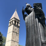 Tall statue of Gregory of Nin reading from a document with a bell tower looming in the background