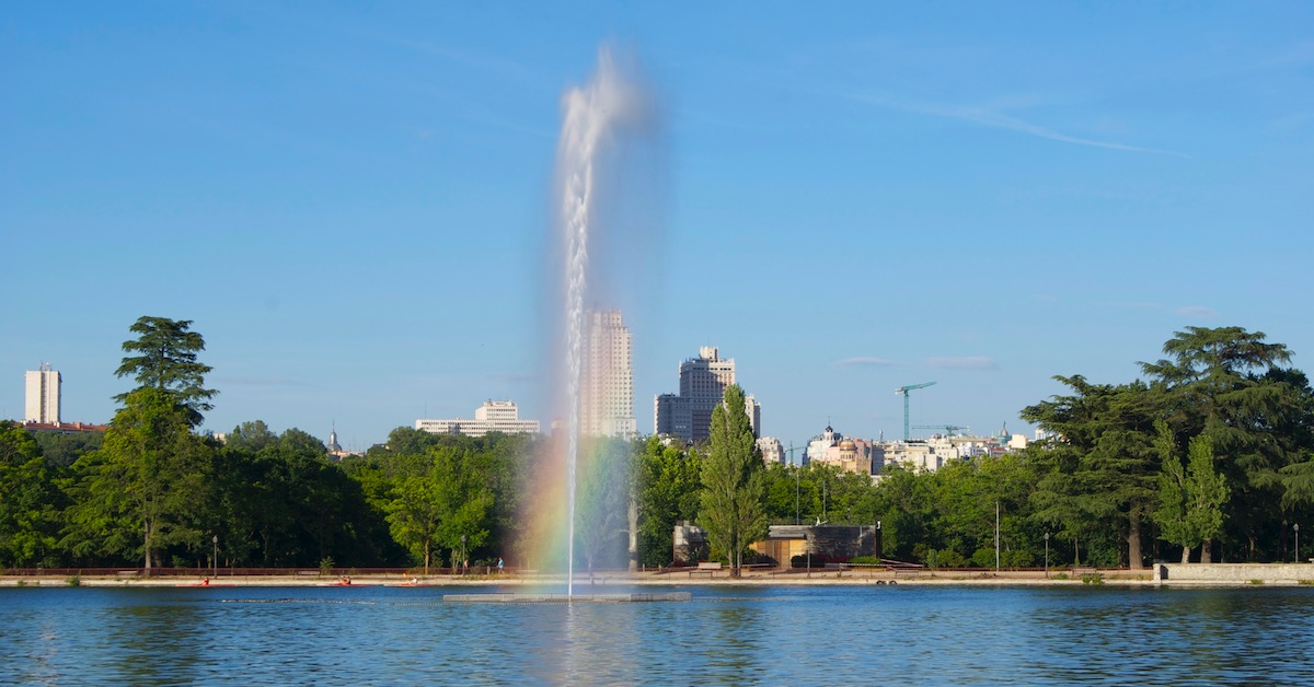 Fountain of water sprays high up from Casa de Campo’s lake with Madrid buildings peeking up out of trees in the background