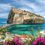 Bright blue flowers in the foreground with the stone buildings of Castello Aragonese surrounded by bright blue water in the middle ground