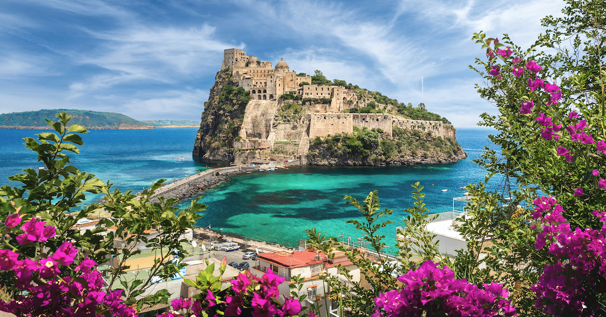 Bright blue flowers in the foreground with the stone buildings of Castello Aragonese surrounded by bright blue water in the middle ground