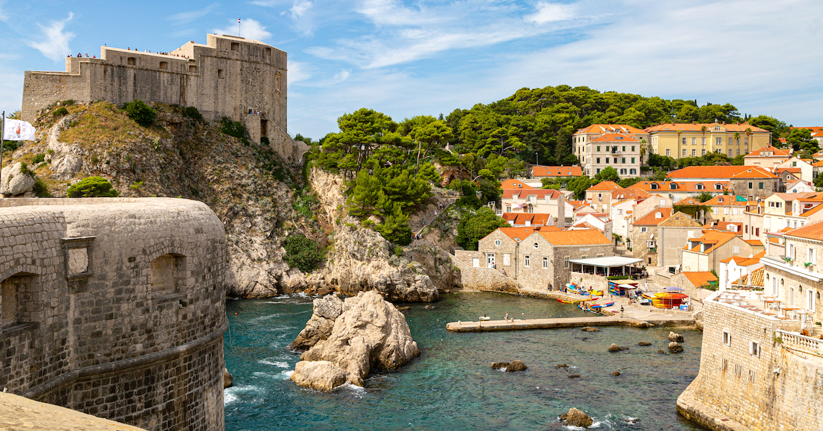 Deep blue bay surrounded by a fortress atop a hill on the left, a rocky green hill in the middle and stone buildings with red roofs on the right in Dubrovnik, one of the most popular Croatia honeymoon destinations