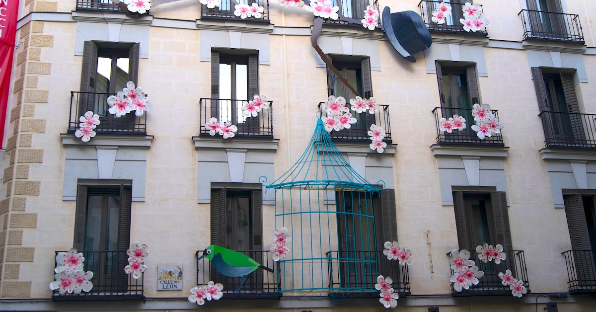 Decorations of a bird, a bird cage, and pink flowers hang from balconies of the same building in Las Letras, Madrid