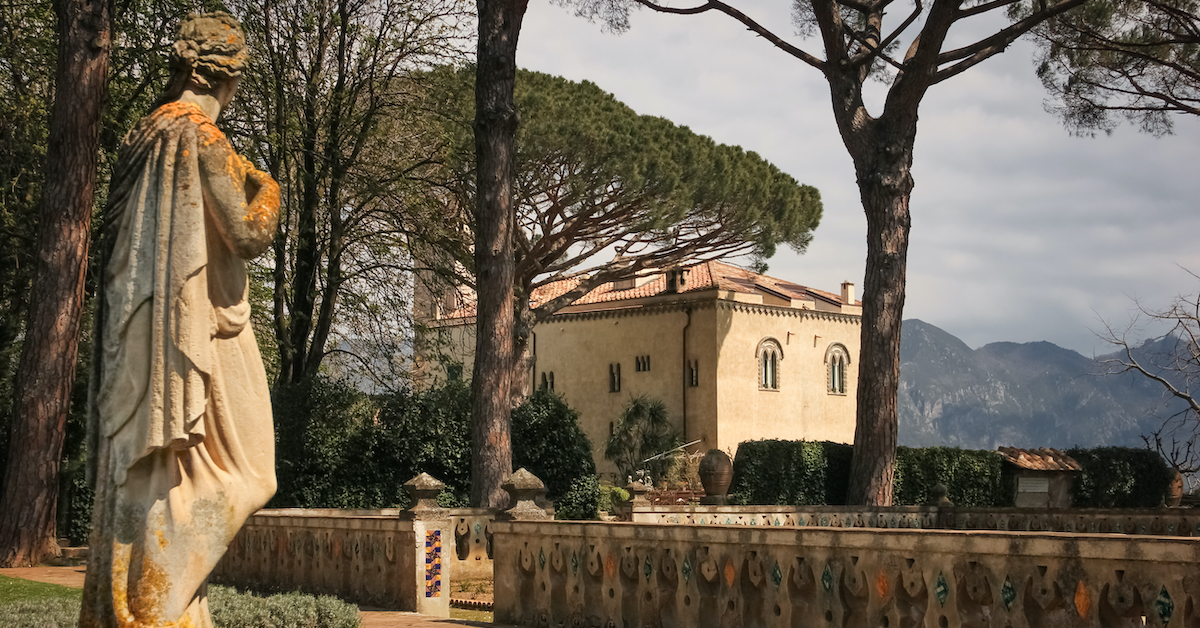 Rear view of a statue of a woman on the left with a beige stone house, trees, and mountains in the background of the Villa Cimbrone Gardens