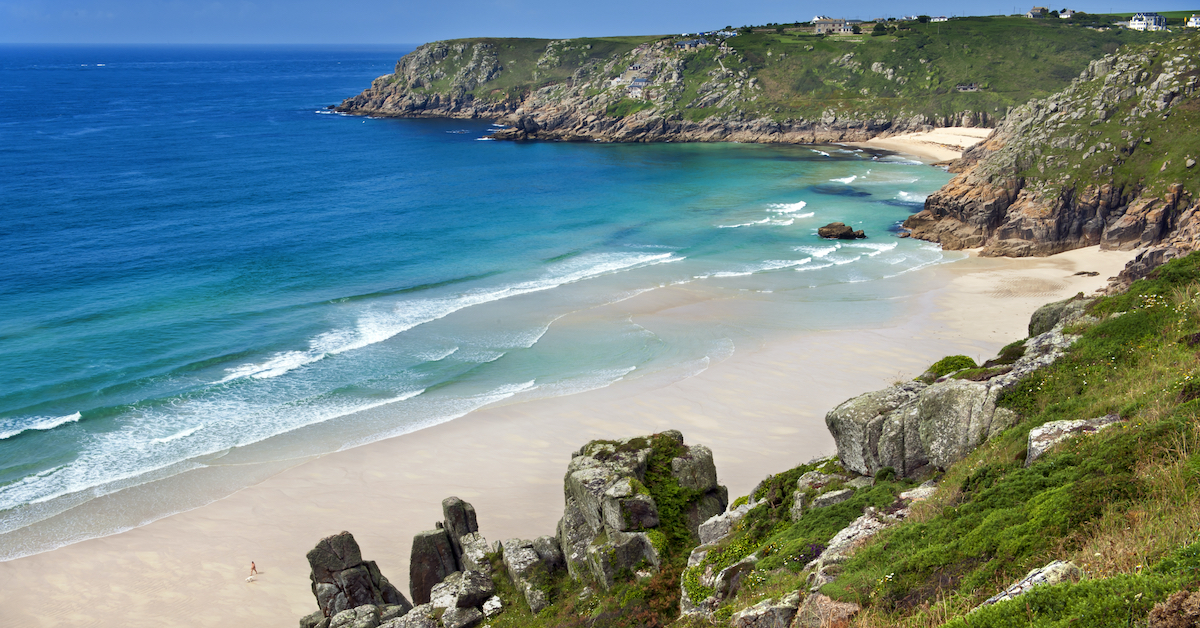 Grassy rocks surrounding the beige sand and blue ocean of Porthcurno Beach in the United Kingdom