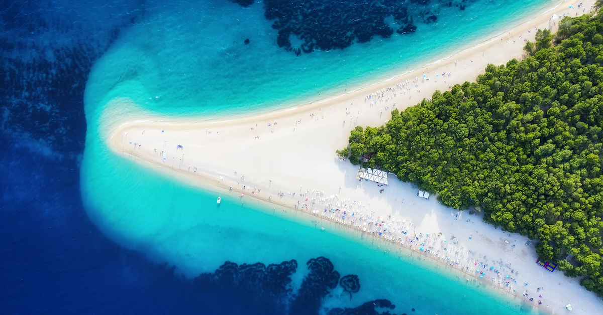 V-shaped white sand beach of Zlatni Rat, one of the best beaches in Europe, jutting out from the right side, speckled with green trees and beach chair and umbrellas leading out toward blue water that gets darker the farther it is from the sand