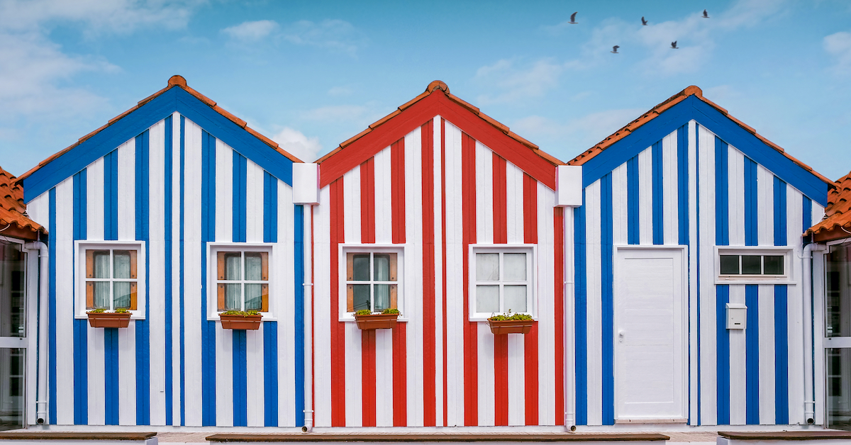 Small red-and-white-striped house in between two small blue-and-white-striped houses with three seagulls flying above them on Portugal's Silver Coast