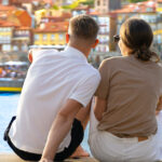 Rear view of a young man and woman sitting on a river bank. The man is pointing towards a boat in the water with colorful buildings in the background