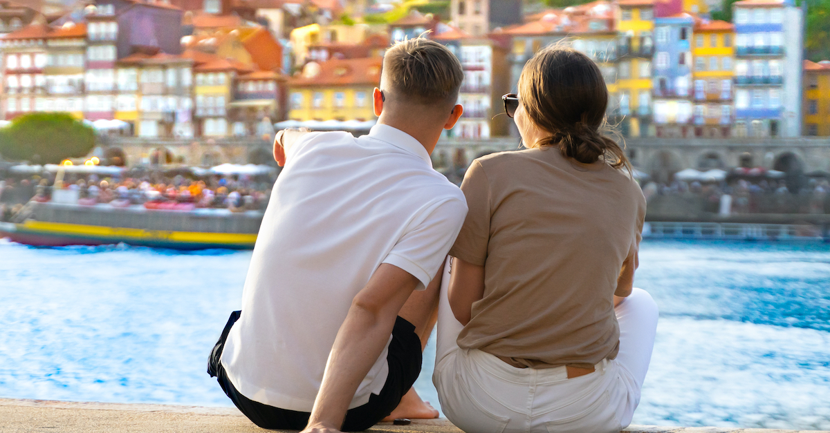 Rear view of a young man and woman sitting on a river bank. The man is pointing towards a boat in the water with colorful buildings in the background