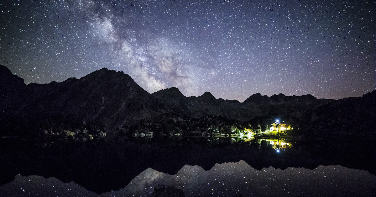Bright starry sky with the Milky Way visible over snow-capped mountains, reflected in a lake in the foreground in the Pyrenees