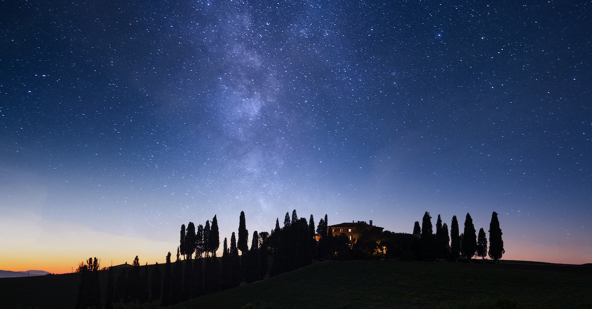 A small stone villa surrounded by cypress trees in Tuscany at night, with the sky lit up by the Milky Way
