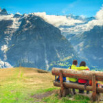 Young couple sitting on a bench overlooking the snowy mountains and green valleys of the Bernese Oberland, one of the best Switzerland honeymoon destinations
