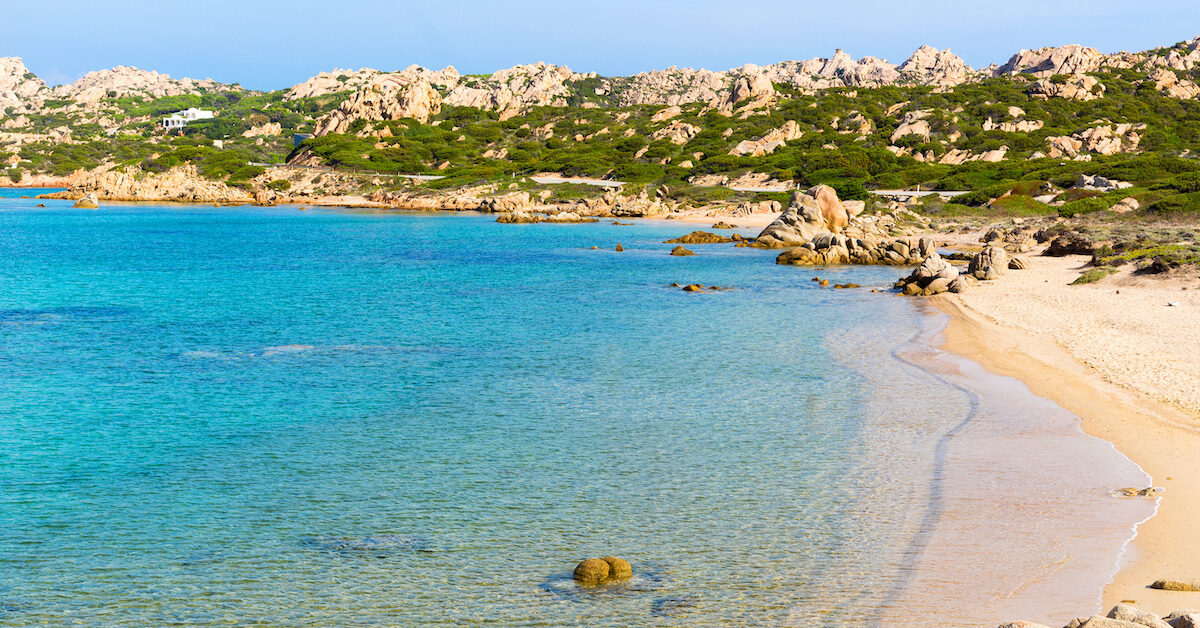 Bright blue water on the left with golden sands on the right and rocky greenery in the background at a beach in Sardinia, one of the most luxurious Italy honeymoon destinations