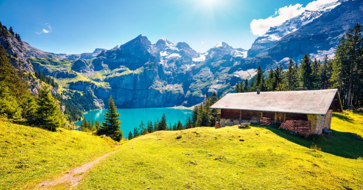 Bright blue lake surrounded by snow-capped mountains with green grass and a wooden cabin leading up to it in Switzerland’s Bernese Oberland