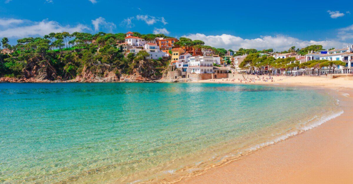 Sandy beach and crystal blue waters with greenery and a colorful town perched on cliffs in the background on Spain’s Costa Brava, one of the most relaxing vacations throughout Europe