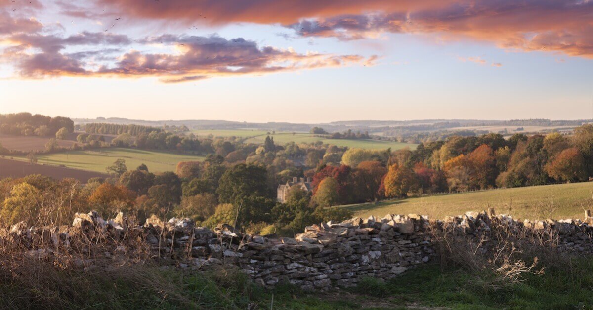 Stone wall in the foreground with rolling green hills, fall foliage and quaint stone buildings of the Cotswolds in the background