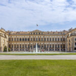 A sprawling tan and white palace with a fountain in front and an Italian flag on top
