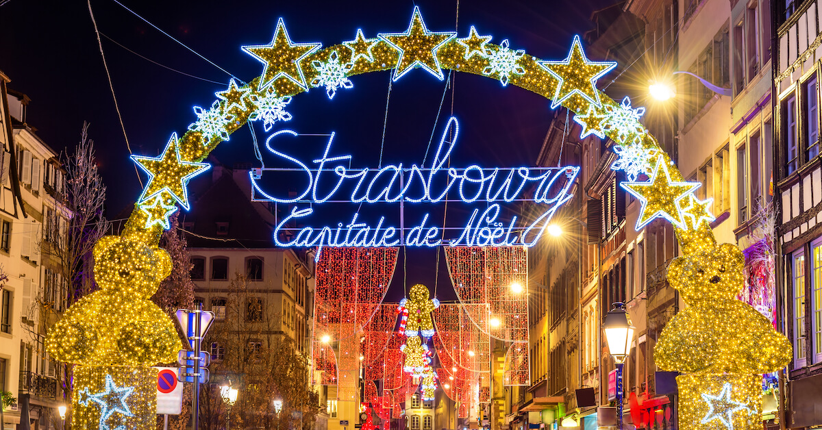 A street lined with arched of holiday lights, the frontmost of which has a sign that reads "Strasbourg: Capitale de Noel"