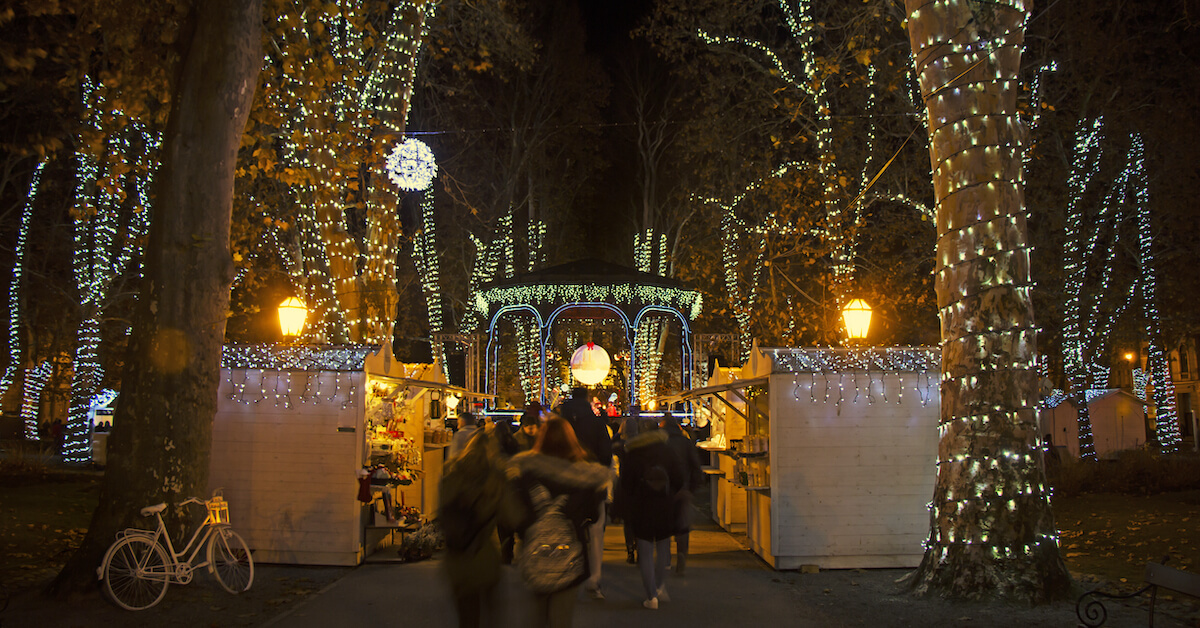 A busy park with market stalls and trees wrapped in lights at night